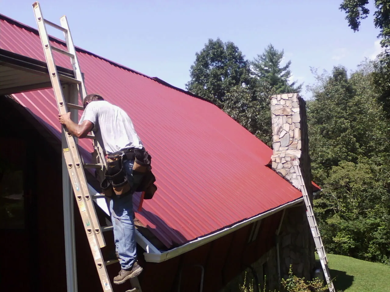 Roofer on a ladder beside a red metal roof and stone chimney — Soffit & Fascia Repair by Roofing Brothers in Latonia